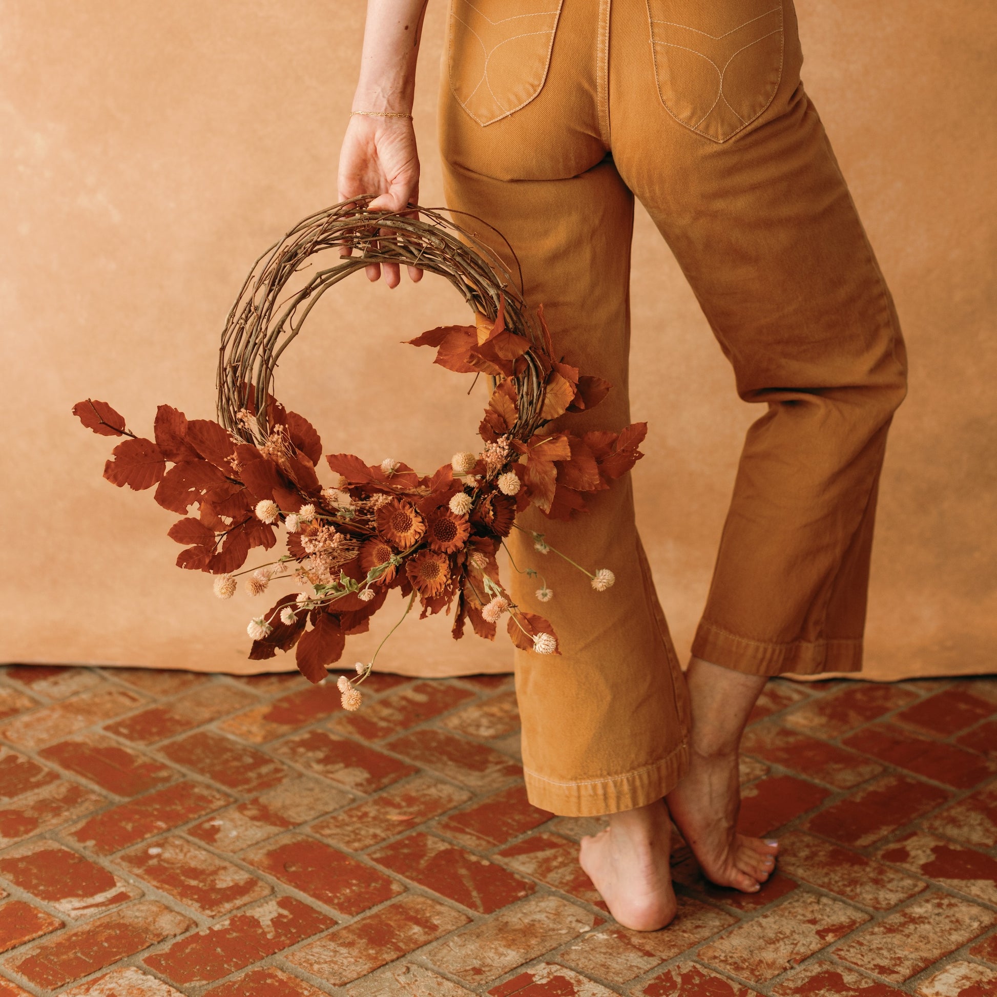 Fall wreath made with rust colored leaves and dried flowers held by woman in ochre pants